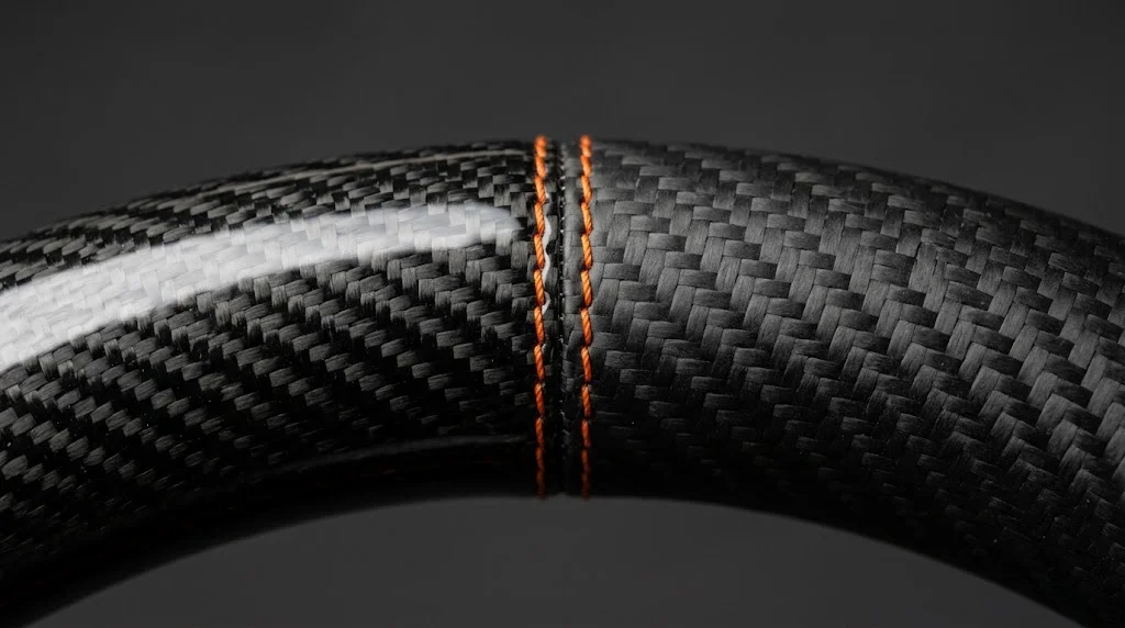 Close-up comparing high-gloss carbon fiber and matte carbon fiber on a BRONFELD Ford steering wheel rim with orange stitching