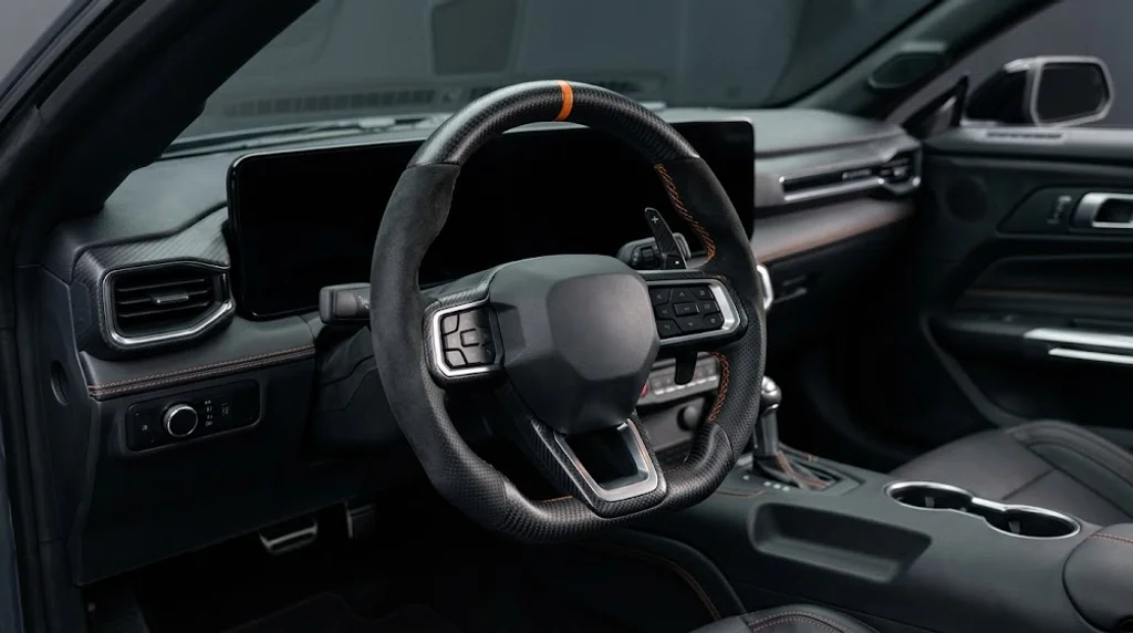 Interior view of a BRONFELD custom steering wheel for Ford Mustang combining three materials: matte carbon fiber trim, black Alcantara upper grips and perforated black leather lower grips with orange stitching